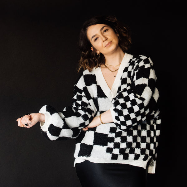 A woman posing in a black and white checkered knit cardigan sweater and a black satin skirt against a solid black studio background.